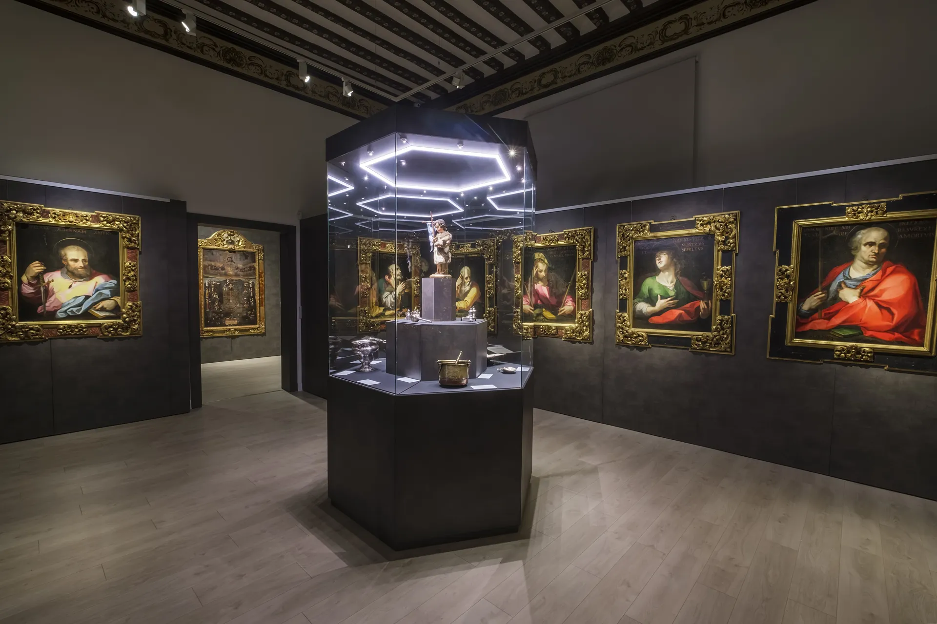 Sala del Apostolado del Palacio Episcopal de Segovia, con cuadros de apóstoles enmarcados en pan de oro y esculturas litúrgicas expuestas en vitrinas.