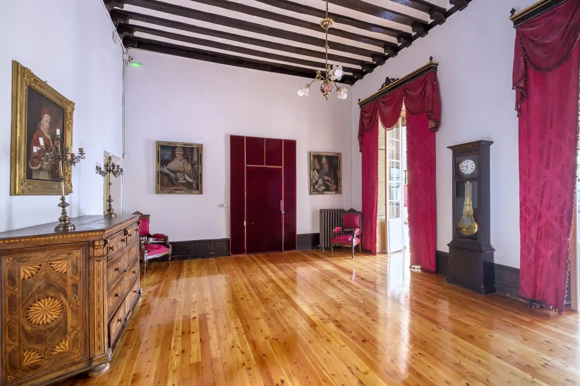 Sala de Espera del Palacio Episcopal de Segovia con cómoda de marquetería del siglo XVIII, retratos de figuras eclesiásticas y cortinas de terciopelo rojo.